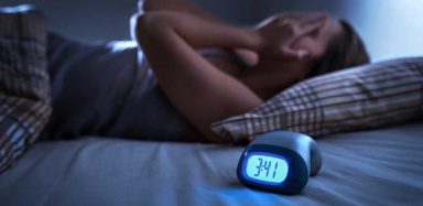A person laying in bed at night with their hands on their face with an alarm clock next to them.